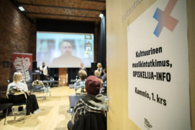 Opiskelijat istuvat luokassa. Kuva on otettu ovelta päin ja auki olevassa on ohjelappu tilaisuudesta.