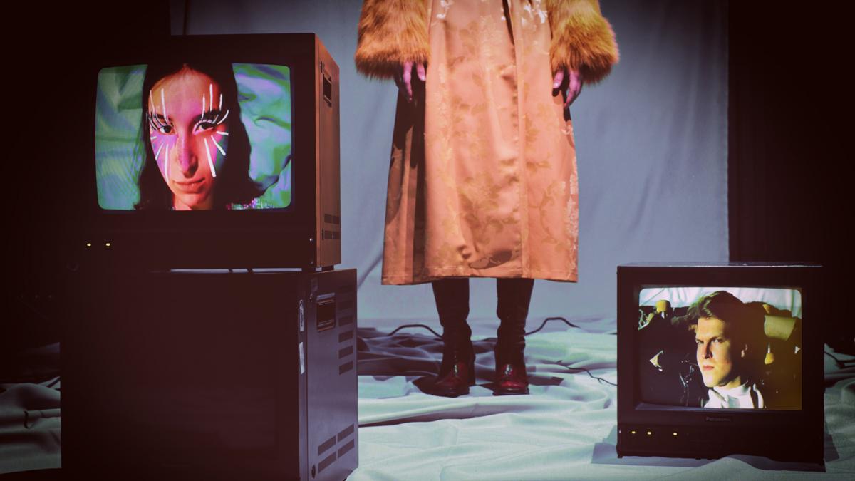 Kuvassa kaksi tv:tä ja henkilö, joka näkyy vyötäröstä alaspäin.