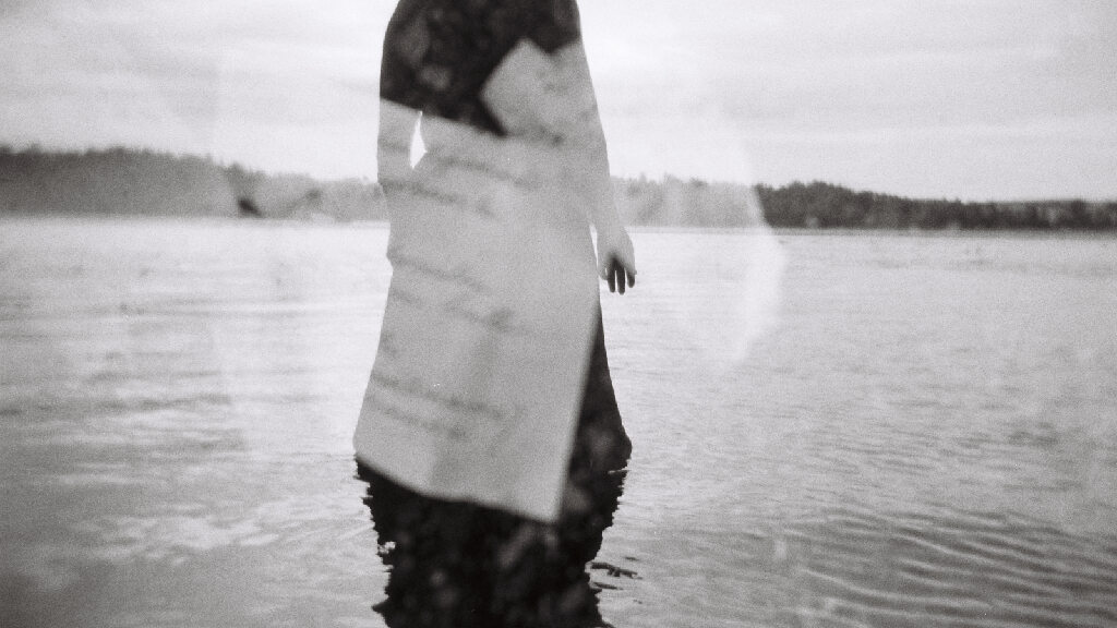 A black and white photo of a woman's body in dark clothes cropped without the head. Behind her a large body of water and the tree line. White pieces of paper with some writing are reflected on the image.