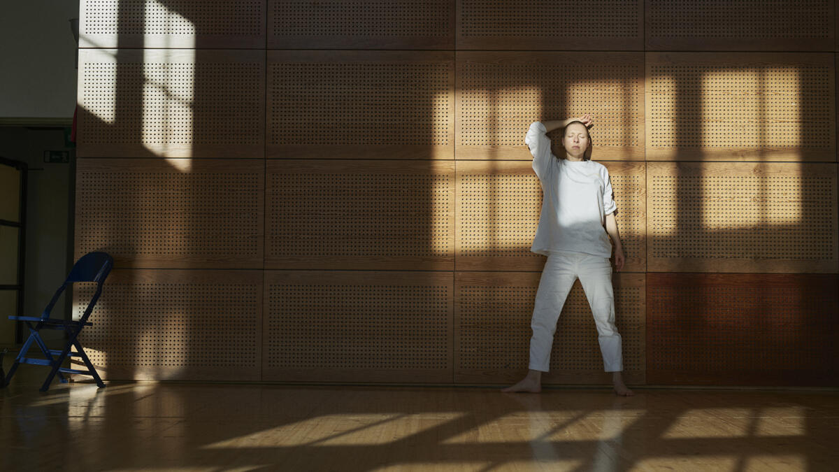 Tanssija valkoisissa vaatteissa nojaa seinään Teatterikorkeakoulun tanssisalissa. Ikkunan varjo heijastuu seinälle ja tanssijan päälle.