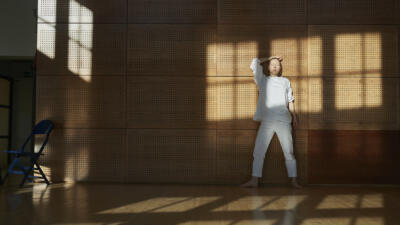 Tanssija valkoisissa vaatteissa nojaa seinään Teatterikorkeakoulun tanssisalissa. Ikkunan varjo heijastuu seinälle ja tanssijan päälle.