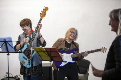 Kaksi eläkeikäistä naista soittaa sähkökitaroita tai -bassoja. Kaikilla on iloiset ilmeet.
