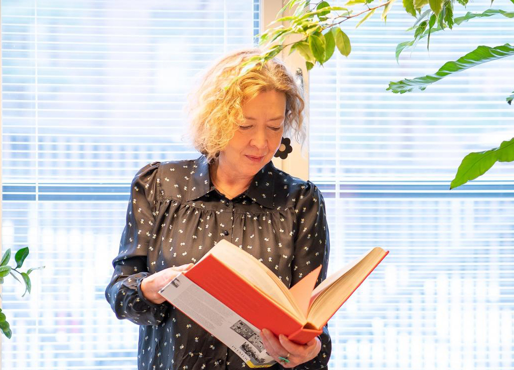 Kuva professori Riikka Stewenistä lukemassa kirjaa ikkunan edessä.