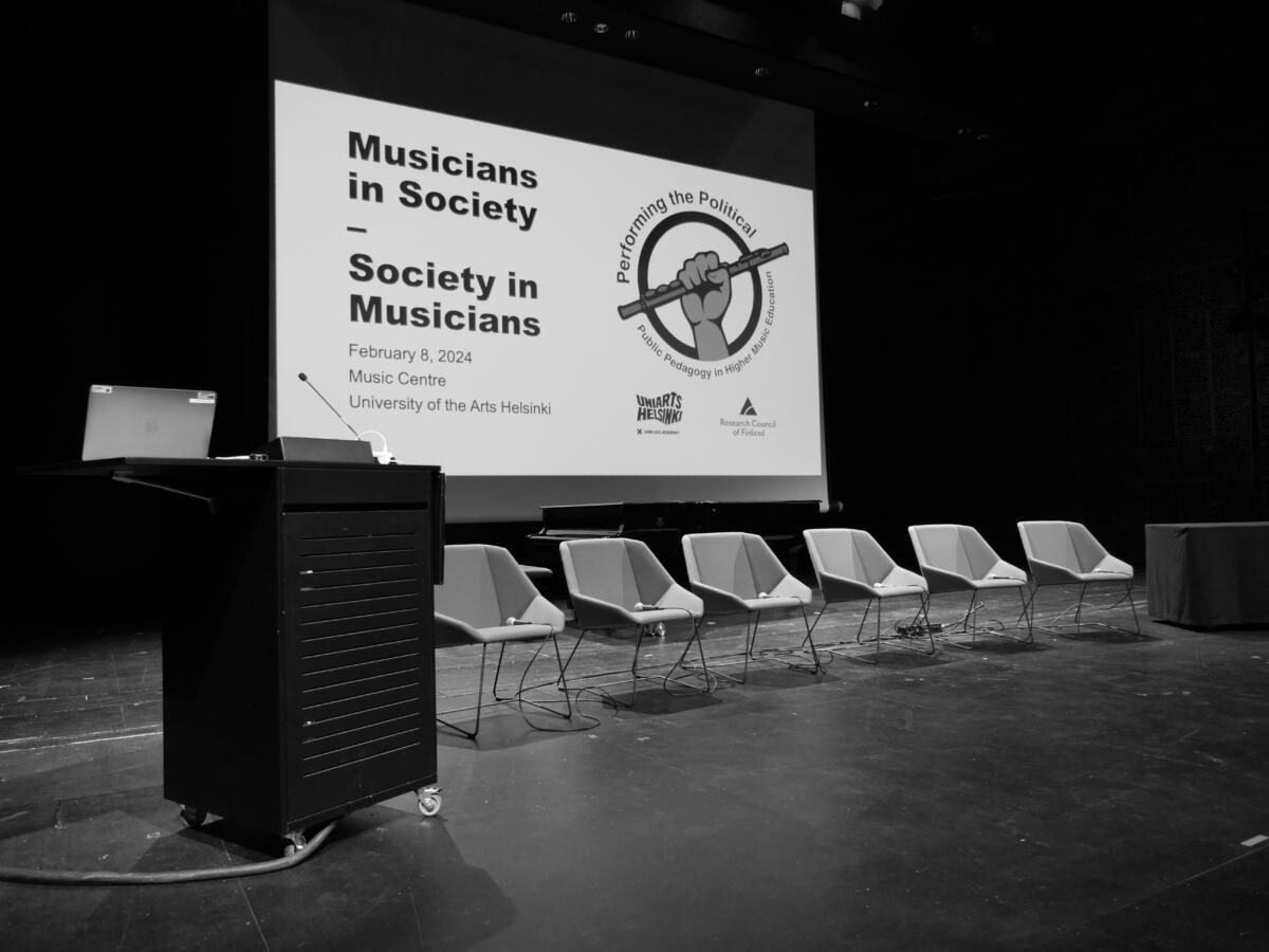 An empty stage with seats on the row and a speaker's podium. Behind the chairs on the screen is a text "Musicians in Society - Society in Musicians" and Performing the political project logo, a fist holding a flute inside a circle.